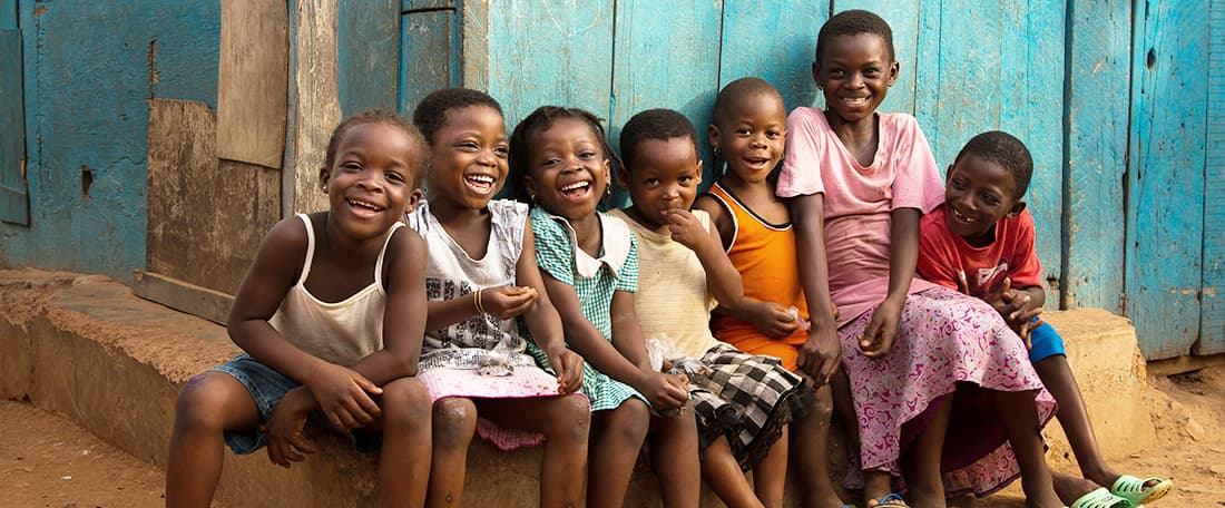 A group of children, young boys and girls, possibly brothers and sisters, siblings, sit together closely in a large group of seven kids, smiling, happy, laughing, outside a poor, low income, poverty, falling apart, peeling old painted blue house, shack, home, boarded building.