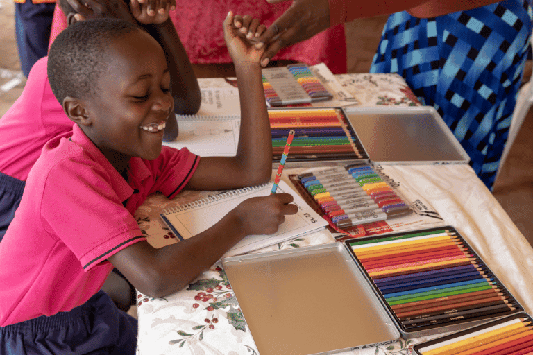 Sarah, 6 years old, Uganda, drawing to help Compassion create new artwork. Sarah wears a pink polo shirt. There are lots of colouring pens and pencils on her table. She is smiling.