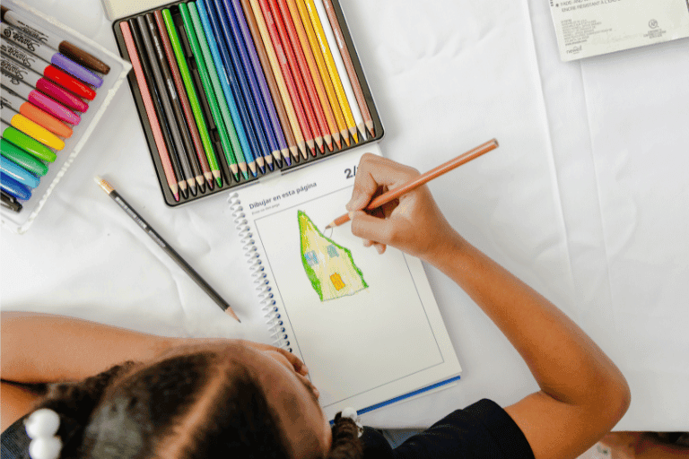 Iriana, a 6 year old girl from the Dominican Republic, draws an illustration of a house. There are coloured pencils in the image, and Iriana's drawing of a yellow house with a green roof.