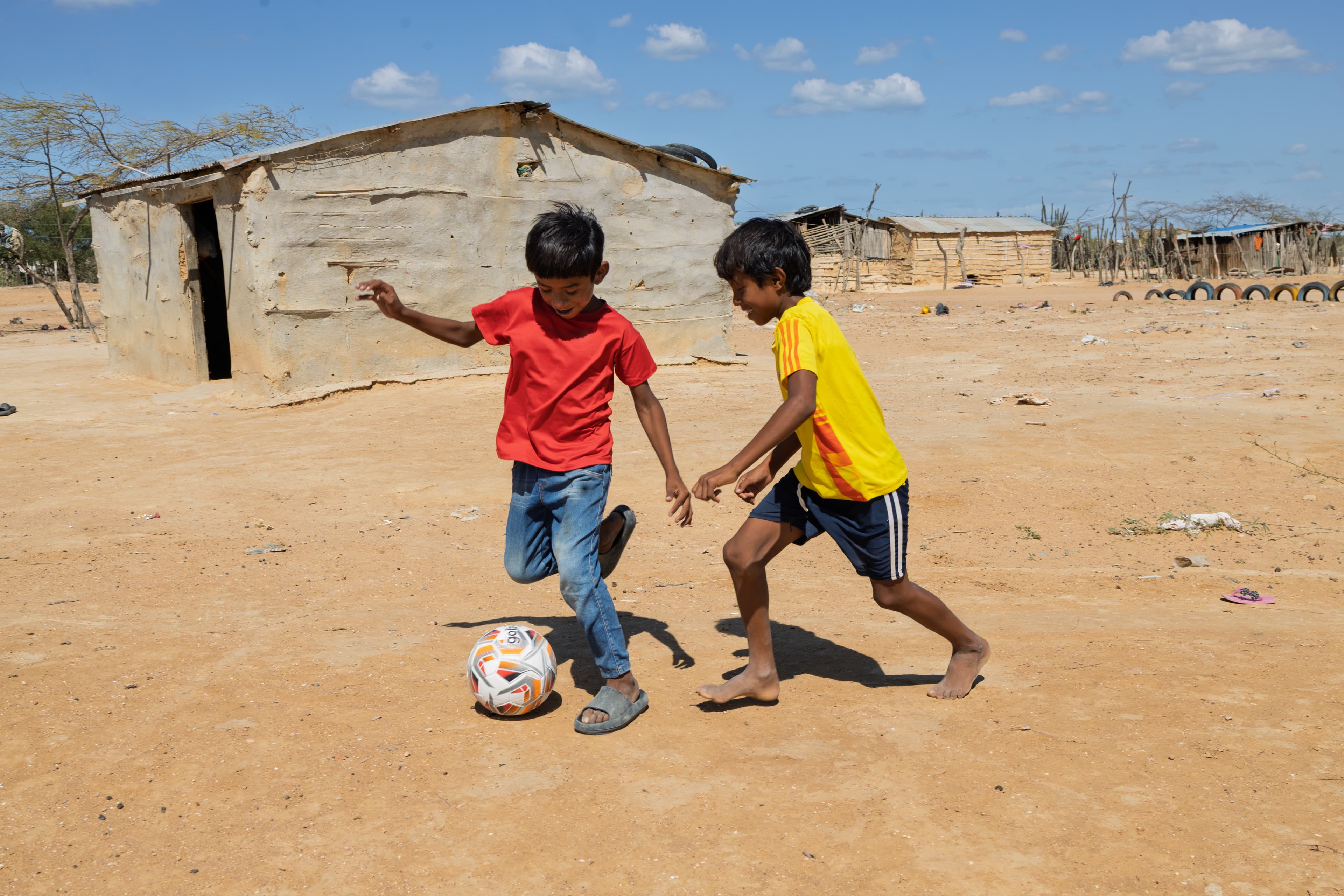 Two boys are playing football on sandy ground in Colombia. Behind them is a house with a corrugated iron roof and a doorway. One boy has a red t-shirt, the other has a yellow t-shirt.