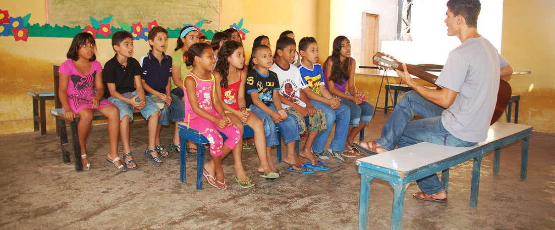 John Nascimento da Silva, a 21 year old, young man, graduate student, wearing a grey shirt and jeans, sits on a small bench inside the classroom, class, yellow painted walls class with colorful mural illustration of family on the back wall, and streamers, playing guitar, music, singing songs, leading music fun class, in front of a group of young centre children, boys and girls, sitting together on benches in three rows, looking to him to lead them in music songs.