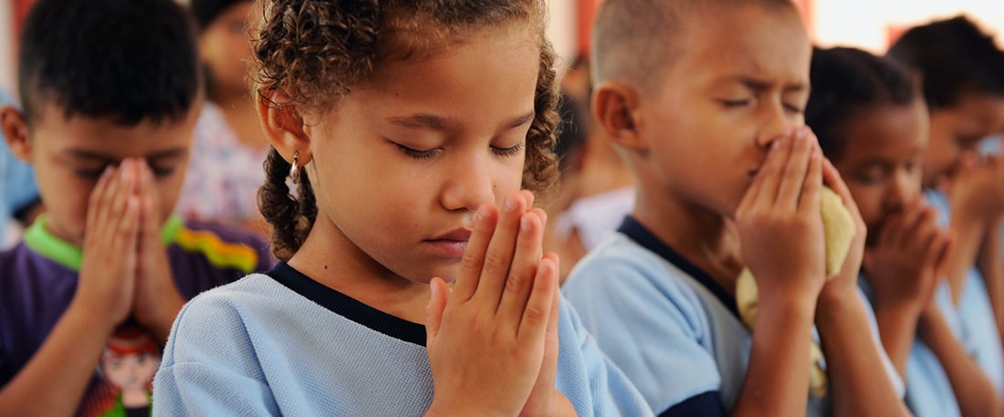 Close up of a project little girl, child, with hands folded, palms held together in prayer in front of her face and eyes closed as she prays. She is praying with other classmates, students, boys and girls, children, inside the project student center classroom.