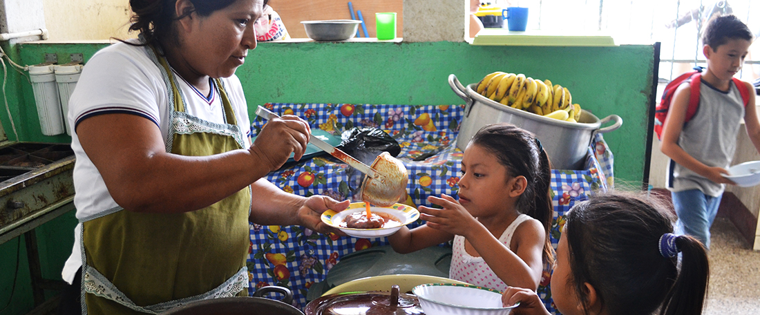 A mother Marta Julia, stands wearing an apron and white shirt, looks down serving plates of food to two girls standing in front of her and the lunch table, as she assists and helps, volunteering in food preparation, cooking, lunch and meal providing at project.