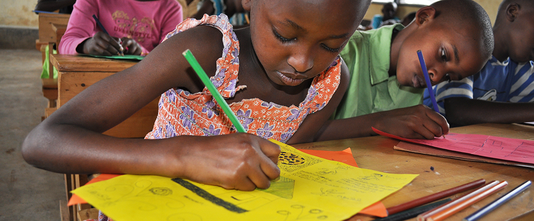 A child, girls, in close-up sits at her desk inside her classroom surrounded by other students, boys and girls, as she looks down drawing, coloring a picture on yellow paper for her sponsor, correspondence. She has a green pencil, coloring pencil in her hand.