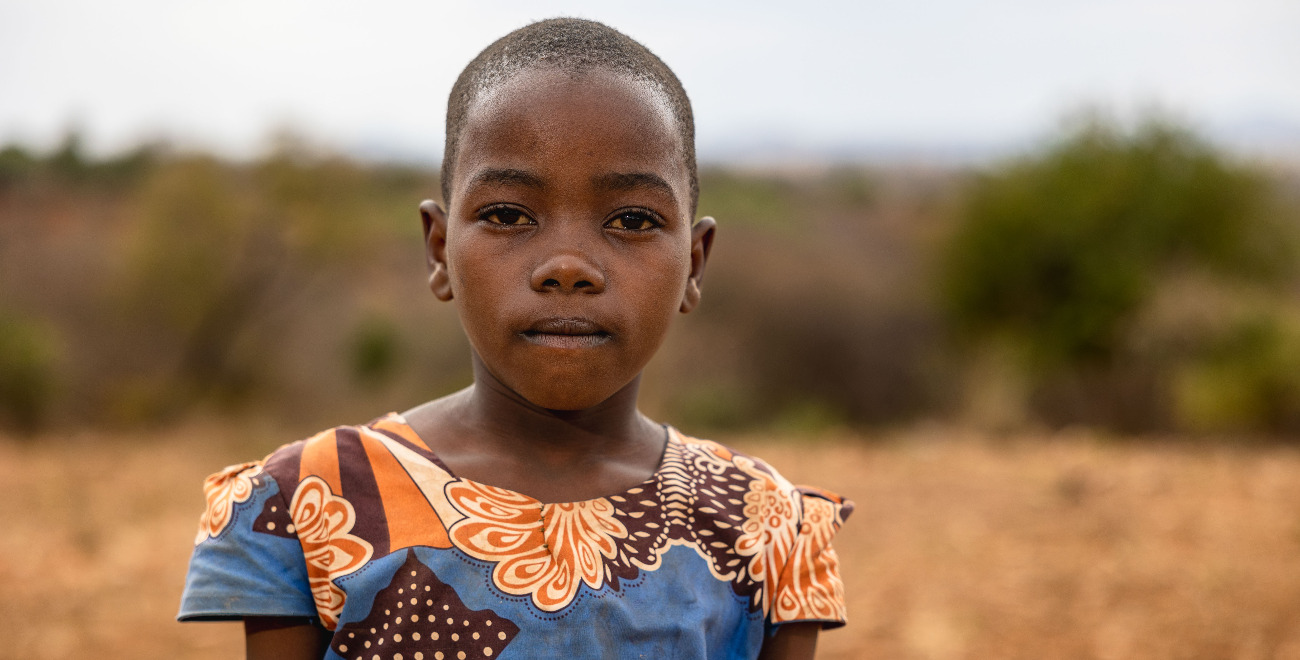 Stella is wearing a blue, brown, and orange patterned dress. She is standing outside her home.