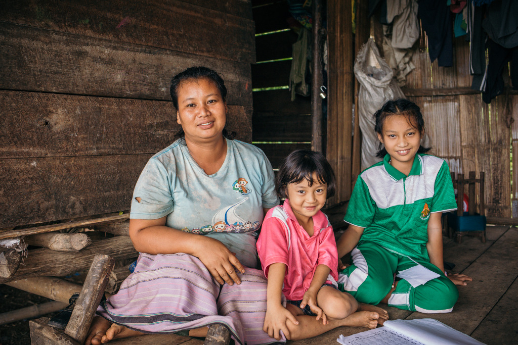 Kamonchanok, wearing a light green shirt and purple skirt, is sitting with her two young daughters, Nirapha, wearing a green and white shirt, and Paphawarin, wearing a pink shirt.