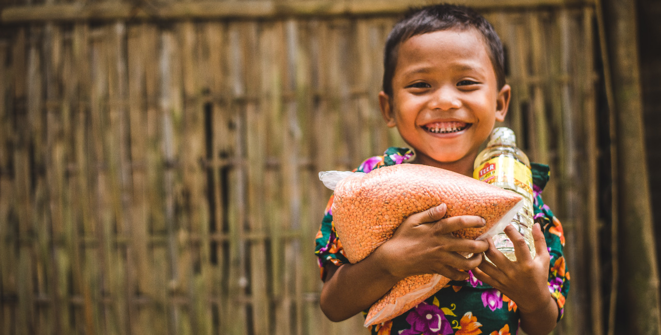 Bhorsha is wearing a green dress with a floral print. She is standing in front of a bamboo wall and is holding food she received from the Compassion centre.